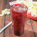 A Carlisle ruby plastic tumbler filled with soda and ice next to popcorn.
