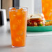 A Carlisle clear plastic tumbler filled with orange liquid and ice on a table with a sandwich.