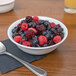 A white GET SuperMel bowl filled with berries on a table with a spoon and a glass of juice.