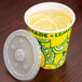 A 16 oz. squat paper lemonade cup with a white plastic lid on a counter with a cup of lemonade and lemon slices.