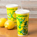 Two 20 oz. tall white paper lemonade cups with yellow and green designs next to two lemons.