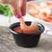A hand dipping shrimp into a Carlisle black plastic ramekin of sauce.