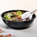 A bowl of salad in a black Cambro Camwear round ribbed bowl with white utensils.
