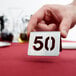 A hand holding a Tablecraft stainless steel number 50 table tent on a white table.