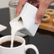 A hand pours milk from an American Metalcraft ceramic milk carton creamer into a cup of coffee.