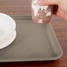 A hand holding a cup over a Cambro rectangular desert tan fiberglass tray on a table in a hospital cafeteria.