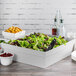 A Tablecraft natural cast aluminum square bowl on a table with a white box of salad inside.