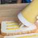 A hand pours yellow mustard from a Vollrath Tri Tip bottle onto a piece of bread.