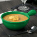 A green Tablecraft cast aluminum soup bowl with a handle filled with soup and croutons with a spoon on the table.