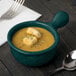 A Tablecraft hunter green cast aluminum soup bowl with a handle filled with soup and croutons with a spoon and fork on the table.