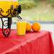 A table with a Creative Converting Classic Red Plastic Tablecover and a drink dispenser with oranges and a glass of orange juice.