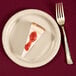 A piece of cheesecake with strawberry jam on an ivory paper plate with a fork.