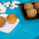A table covered with a turquoise blue Creative Converting tissue/poly table cover with plates of muffins on top.