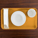 A Cambro rectangular Tuscan gold tray with a bowl and cup on it, with napkins and a sugar packet on the side.