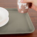 A person holding a cup over a rectangular taupe Cambro tray on a table in a hospital cafeteria.