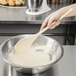 A person stirring white liquid in a bowl with a Thunder Group wooden spoon.