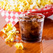 A Fineline clear plastic tumbler filled with soda and ice on a table with a bucket of popcorn.