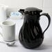 A black Choice Swirl thermal coffee carafe on a table next to a white mug.