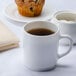 A white Arcoroc stacking mug filled with brown liquid on a table with a muffin.