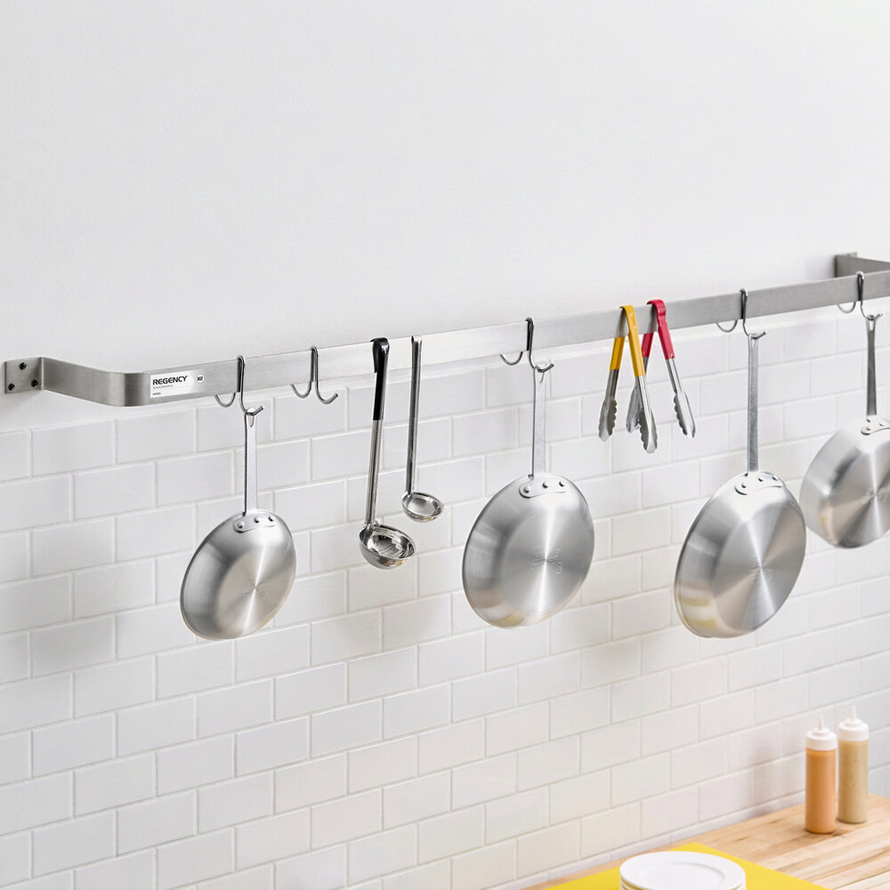 A stainless steel wall-mounted rack with several hooks holding metal pans, ladles, and kitchen tongs against a white tiled backsplash.