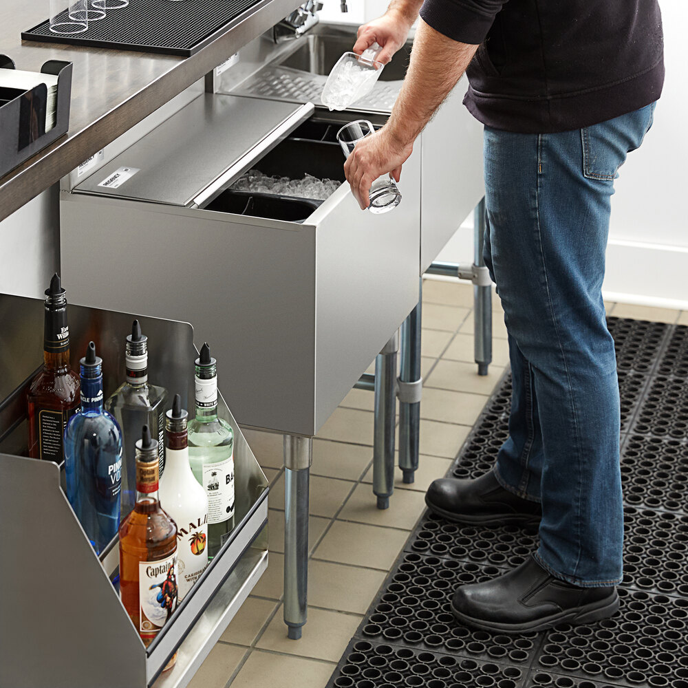 A person is scooping ice from a stainless steel ice bin with a sliding lid in a bar setting, with bottles of liquor visible nearby.