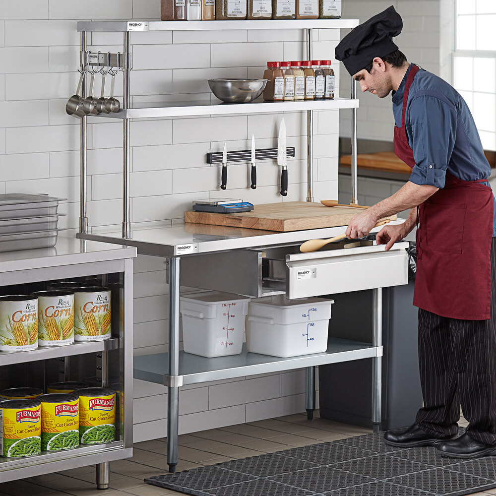 A chef in a commercial kitchen stands at a stainless steel workstation with double deck overshelves holding spices, utensils, and bowls, while opening a drawer beneath the counter.