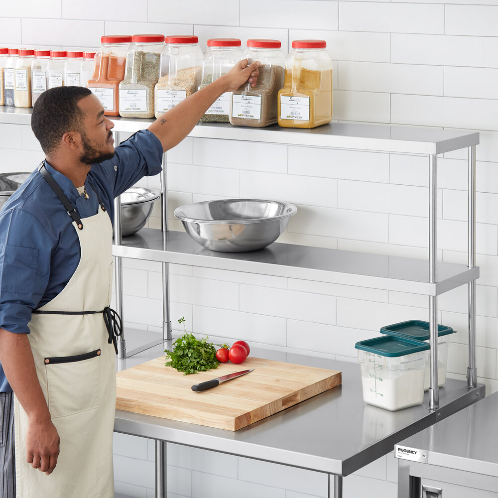 A man in an apron is reaching for a container of spices on the top shelf of a stainless steel double deck overshelf in a commercial kitchen setting.