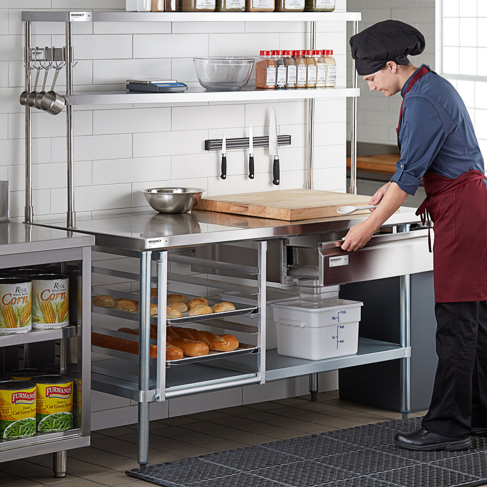 A person in a commercial kitchen is working at a stainless steel prep table with shelves holding bread, spices, and kitchen tools.