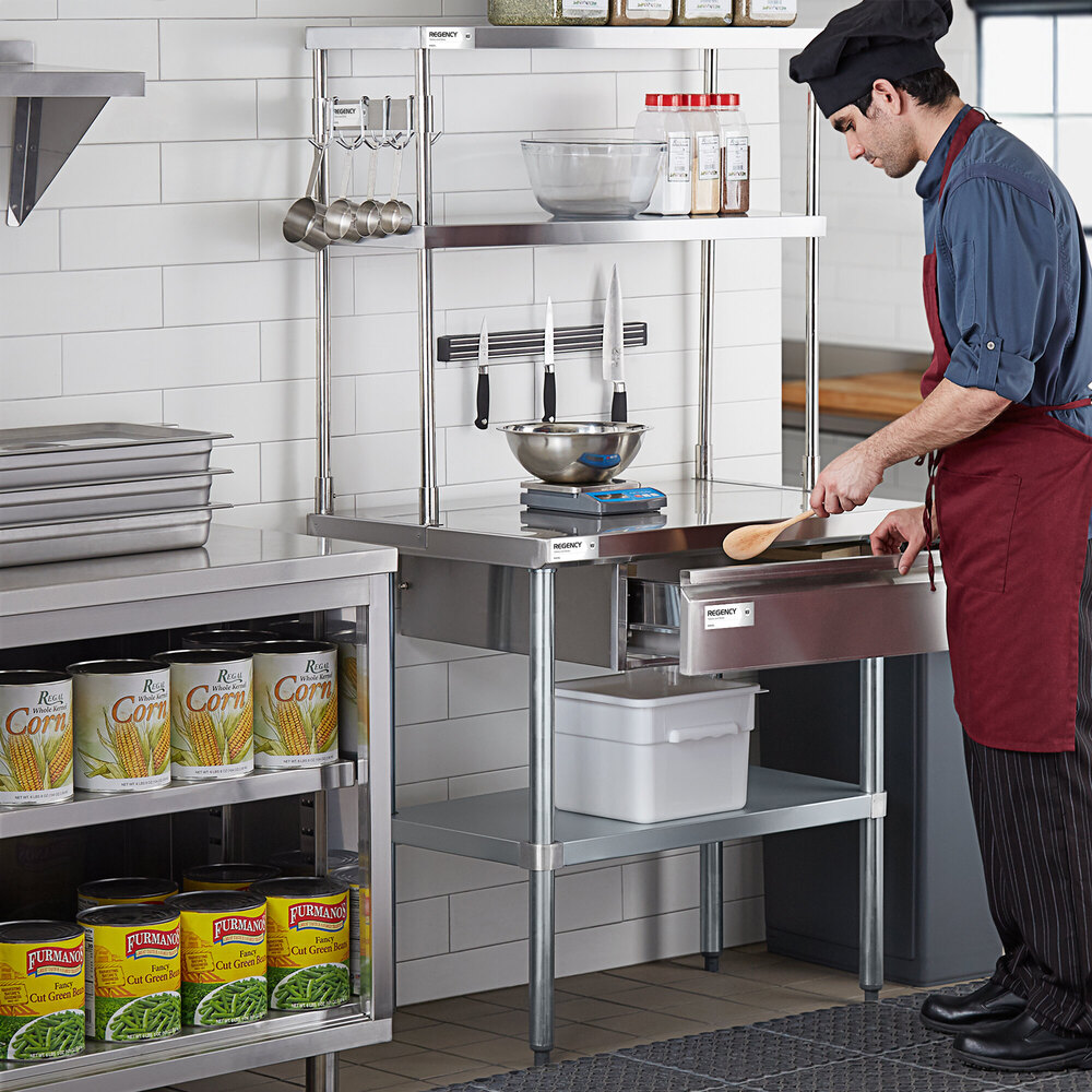 A chef in a commercial kitchen stands at a stainless steel workstation with a double deck overshelf holding bowls, spices, and utensils, with knives mounted on a magnetic strip and canned goods stored below.