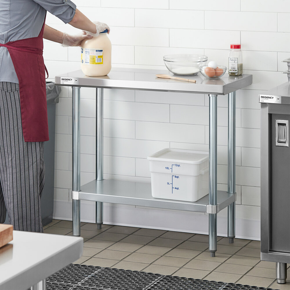 A stainless steel work table with a lower shelf, shown in a kitchen setting with various kitchen items on top and a person working beside it.