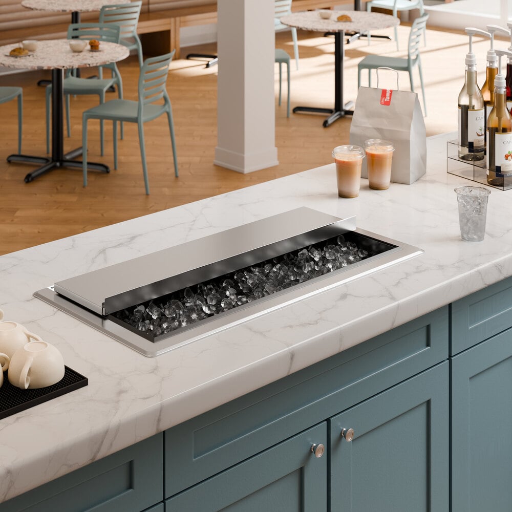 A stainless steel drop-in ice bin with a sliding lid is installed in a marble countertop, partially open to reveal ice inside.