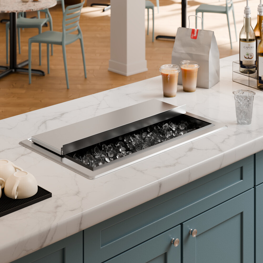 A stainless steel drop-in ice bin with a partially open sliding lid, filled with ice, set into a marble countertop.