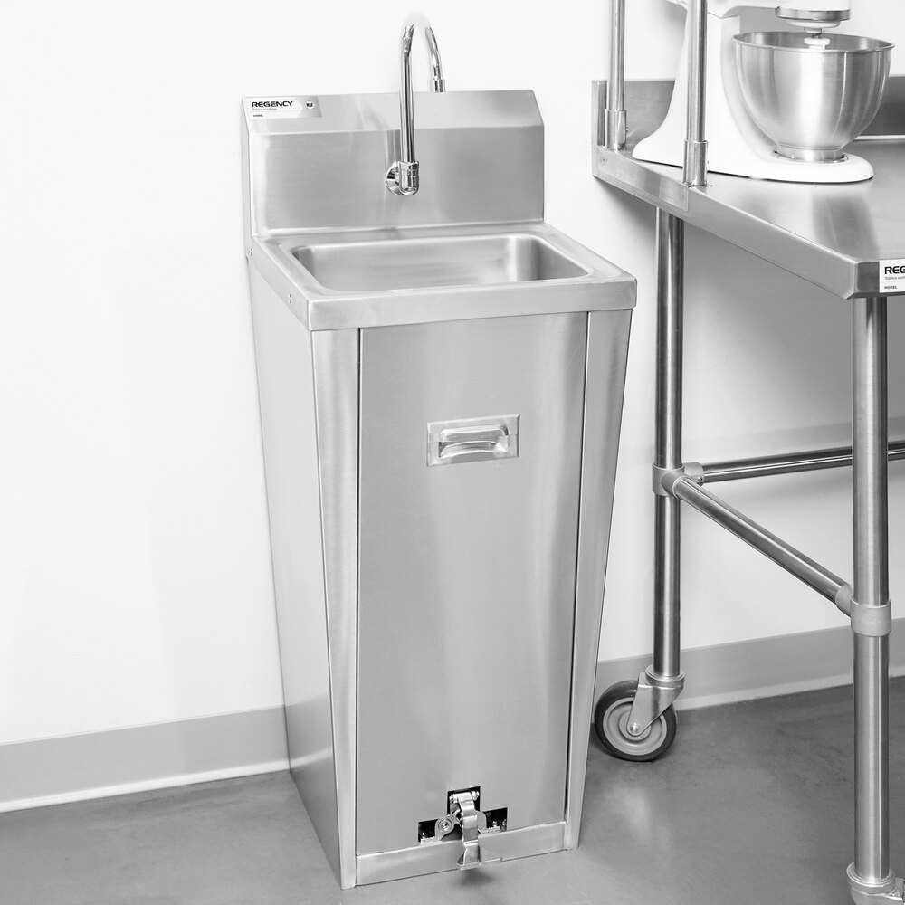 A stainless steel pedestal sink with a tall base, faucet, and foot pedal, positioned next to a metal table in a commercial kitchen setting.