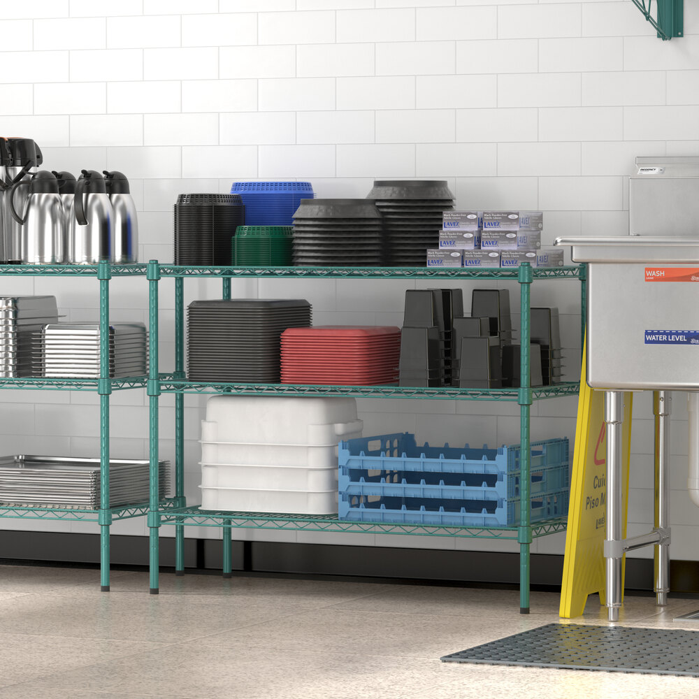 A green metal shelving unit holds various kitchen items such as trays, containers, and pitchers, set against a white tiled wall.