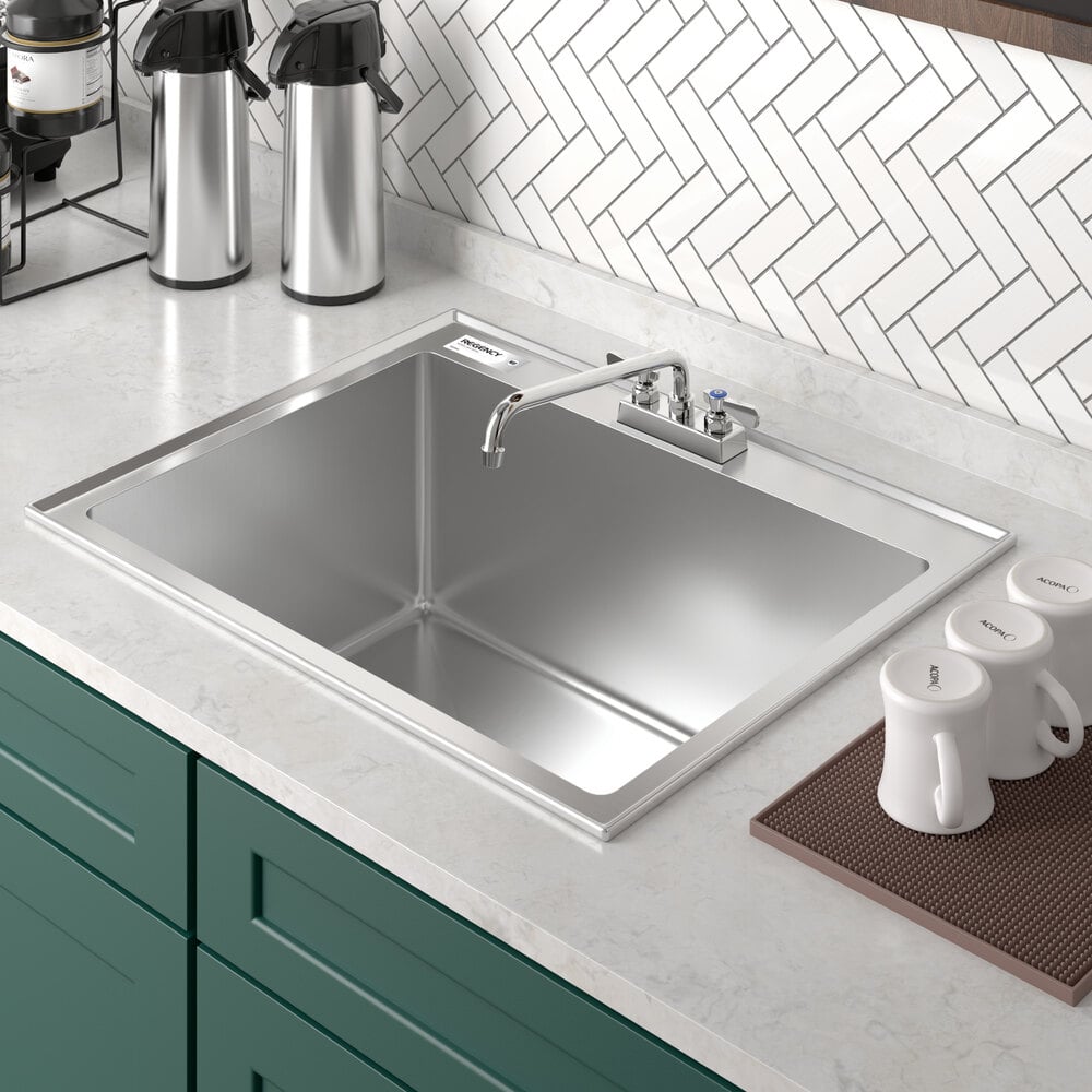 A stainless steel single-basin sink with a faucet, set in a white countertop with green cabinetry and a herringbone tile backsplash.