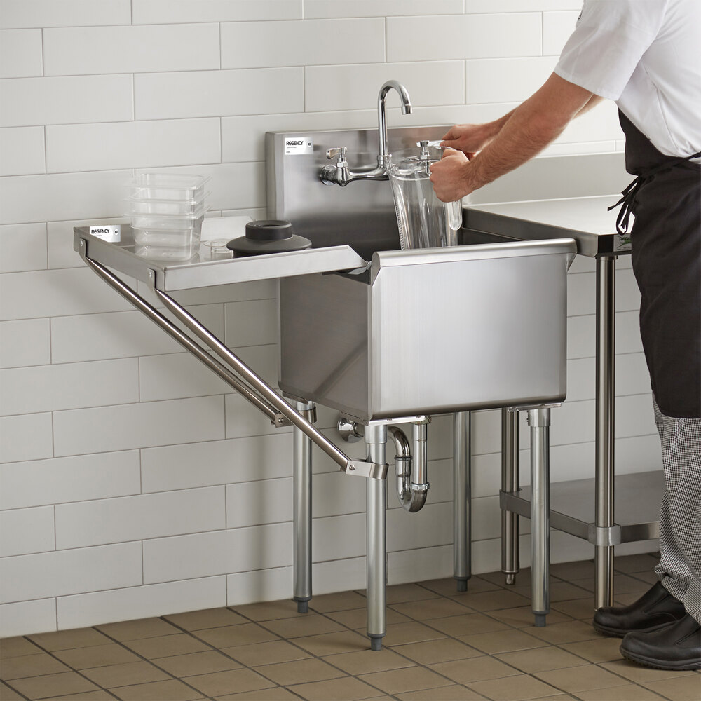 A person is using a stainless steel sink with an attached detachable drainboard in a commercial kitchen setting.