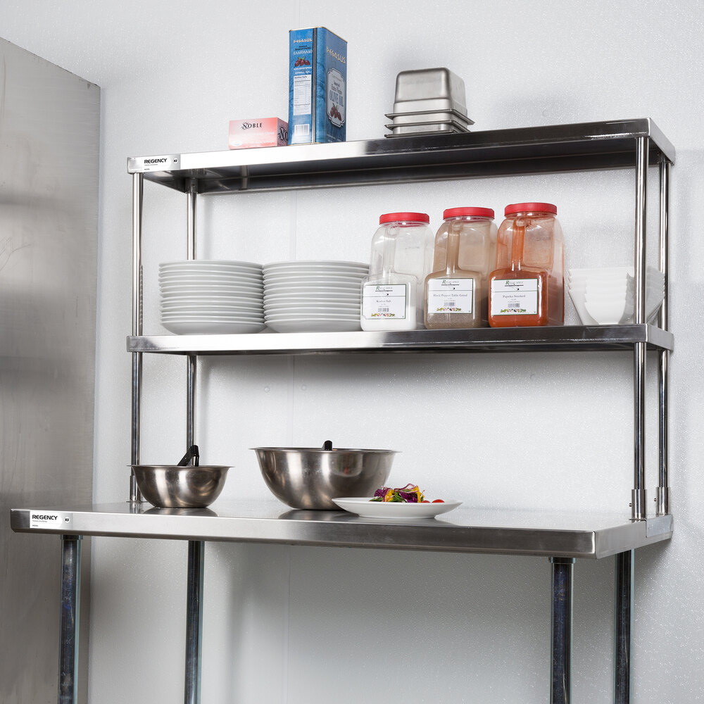 A stainless steel double-deck overshelf mounted above a worktable, holding plates, containers, and kitchen supplies.