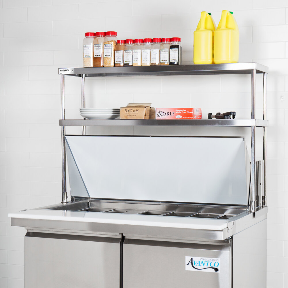 A stainless steel double deck overshelf mounted on a commercial kitchen prep table, holding spices, condiments, and other kitchen supplies.