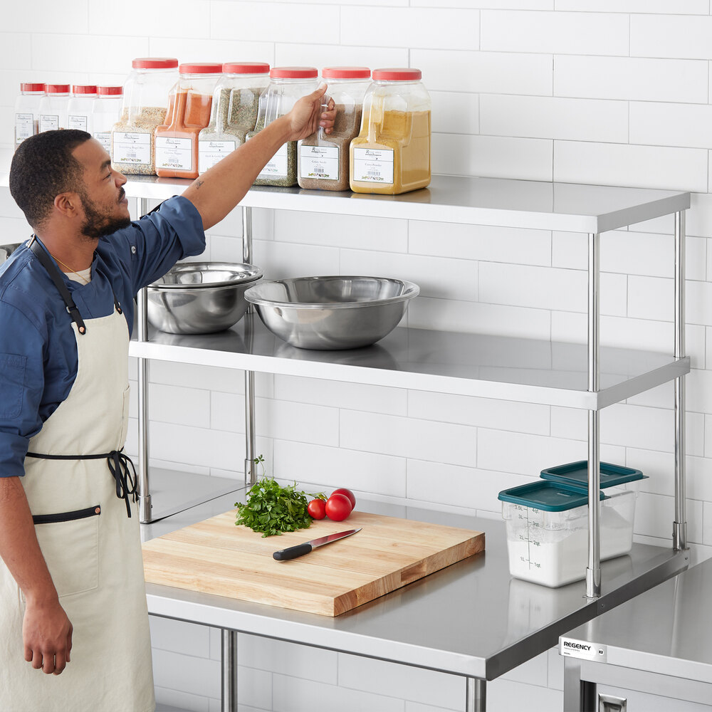 A person in an apron is reaching for a large spice container on the top shelf of a stainless steel double deck overshelf, which also holds mixing bowls, a cutting board with vegetables, and food storage containers.