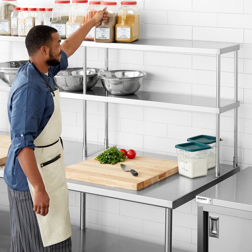 A man in a kitchen reaches for a large spice container on the top shelf of a stainless steel double deck overshelf, with mixing bowls, a cutting board, vegetables, and storage containers visible on the counter below.