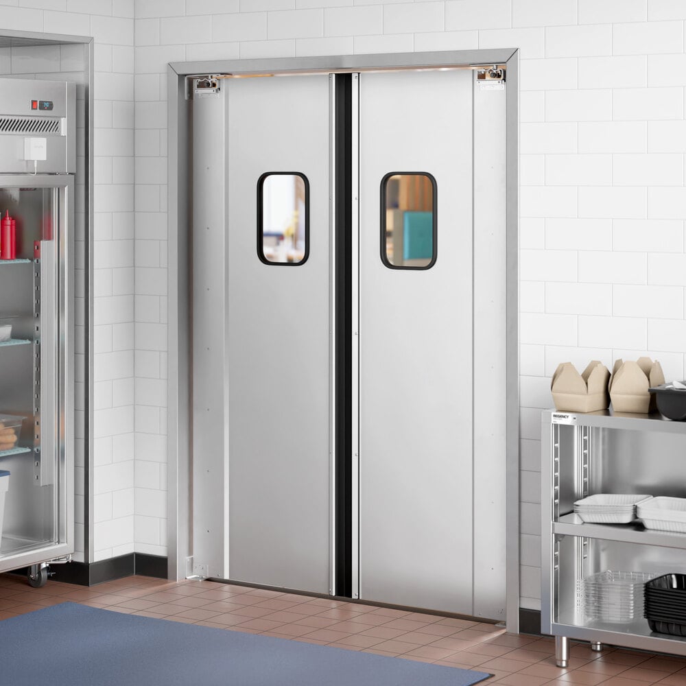 A pair of silver double swinging doors with small rectangular windows, set in a tiled wall between a metal shelving unit and a refrigerator.