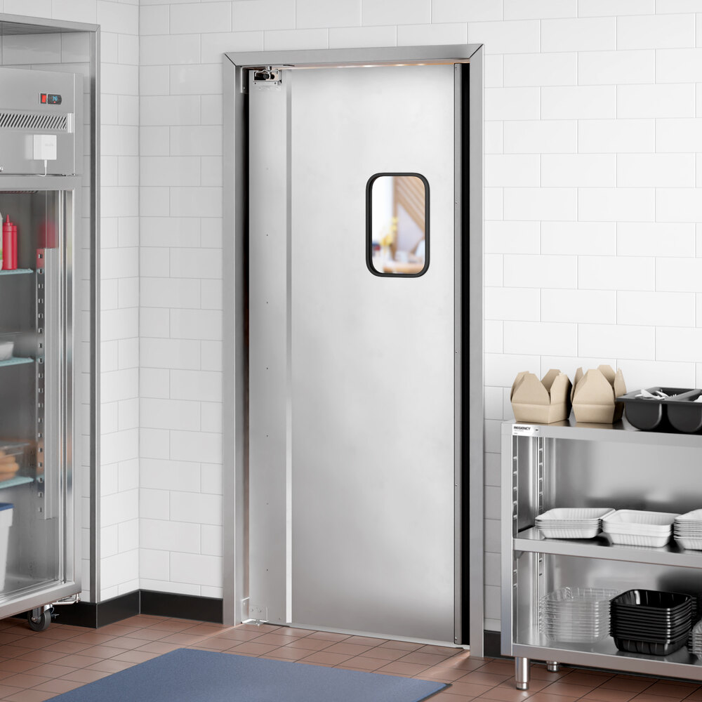 A stainless steel swinging door with a small rectangular window, installed in a commercial kitchen with white tiled walls and metal shelving.