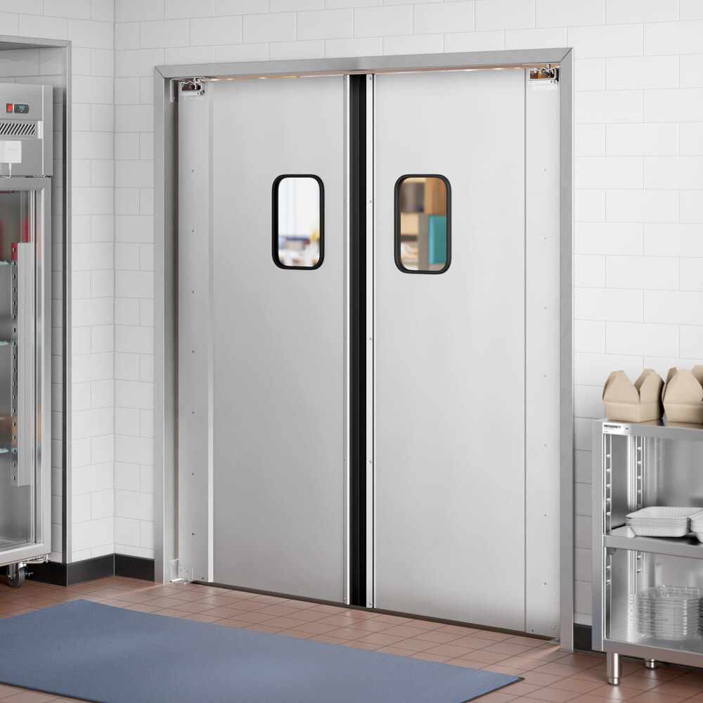 A pair of silver double swinging doors with small rectangular windows, installed in a tiled wall within a commercial kitchen setting.