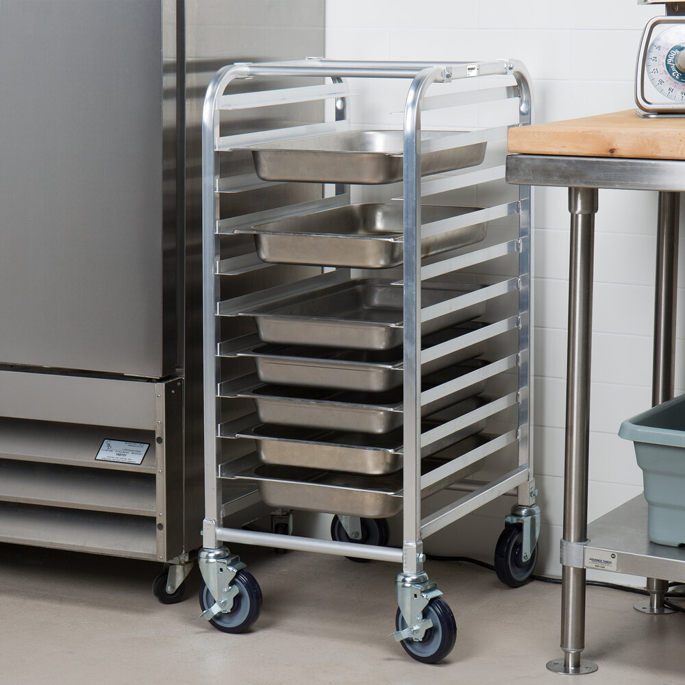 A metal rack on wheels holding several rectangular metal trays, positioned between a refrigerator and a table in a kitchen setting.
