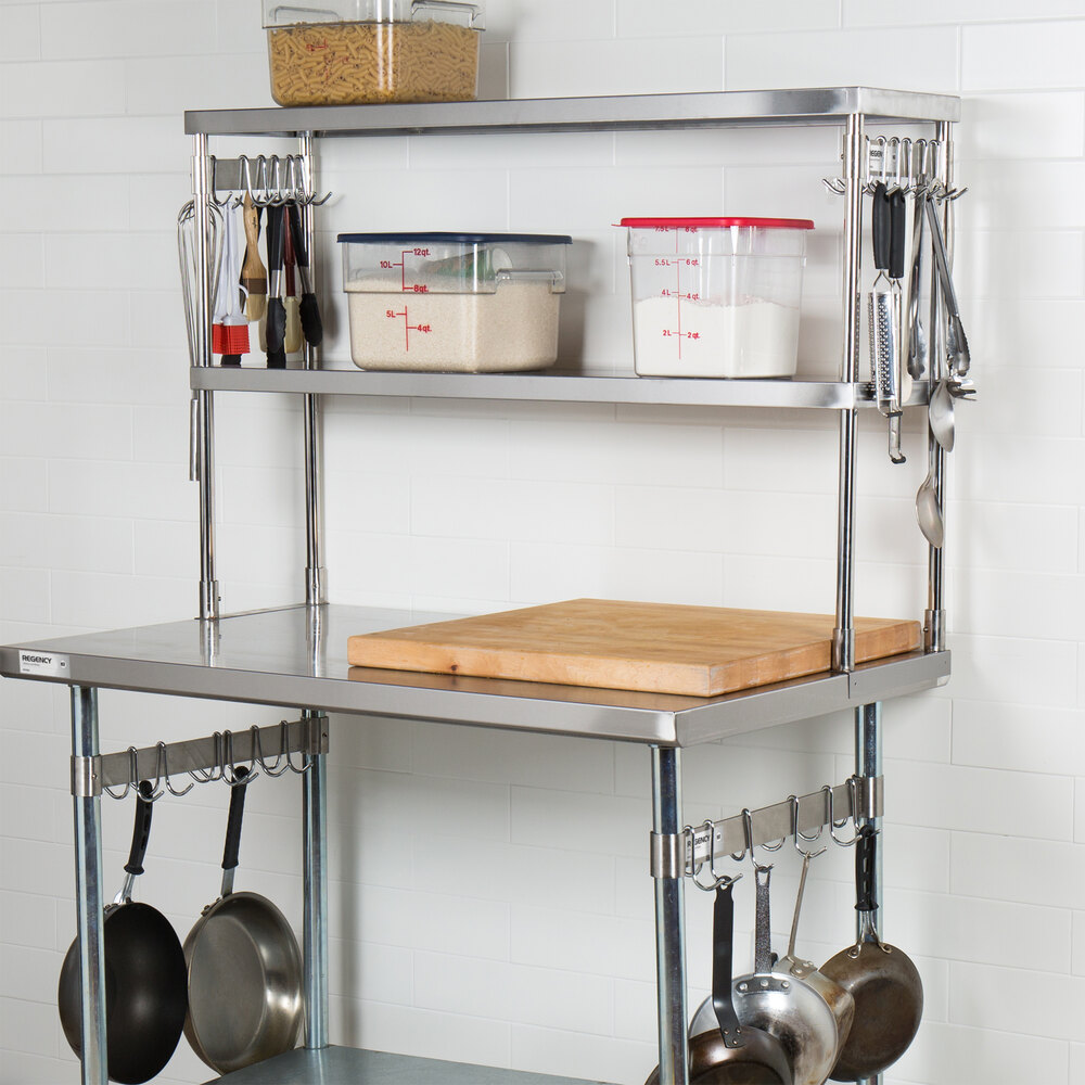 A stainless steel kitchen workstation with an overhead shelf, hanging utensils, pots, and a wooden cutting board on the countertop.