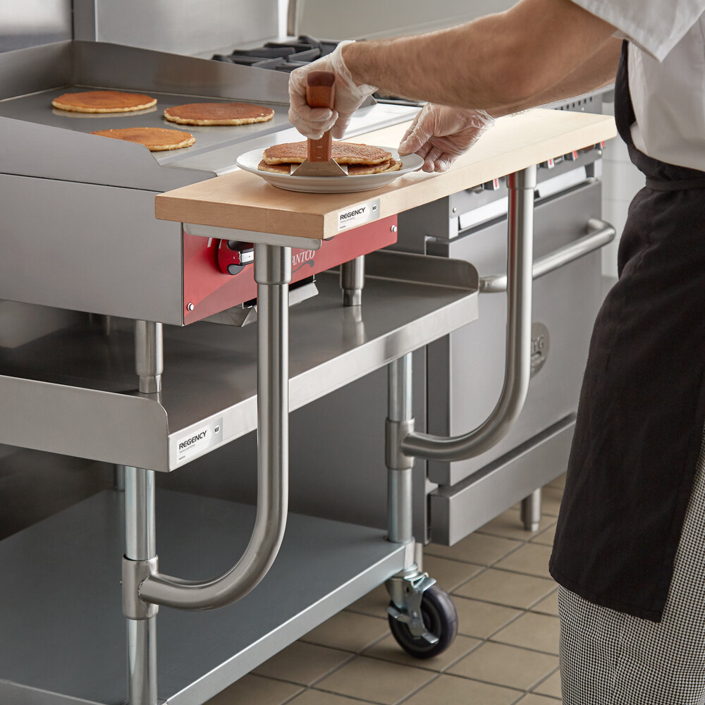 A person is spreading something on pancakes placed on a wooden cutting board attached to a stainless steel kitchen equipment stand.