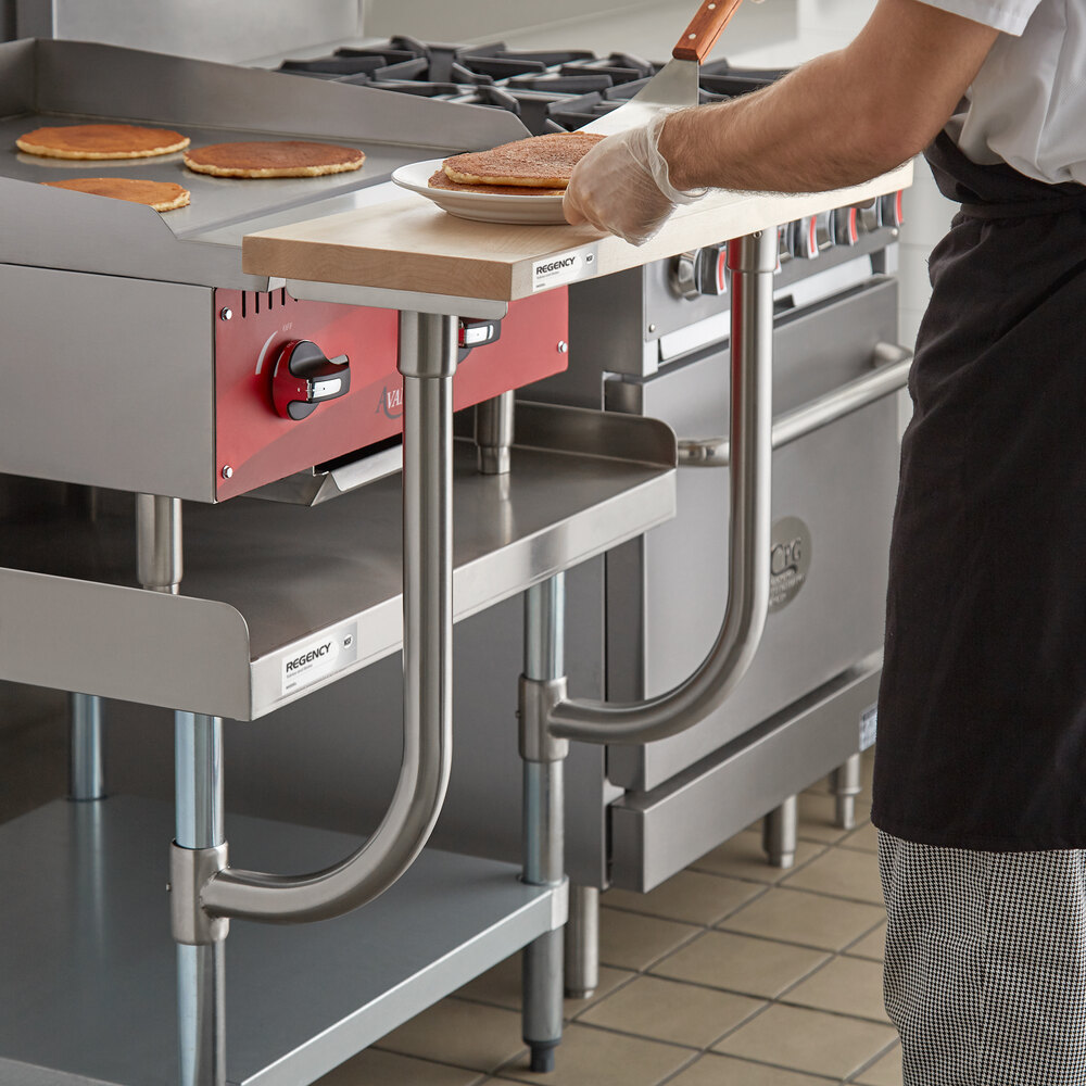 A person is using a wooden cutting board attached to a metal kitchen equipment stand, transferring pancakes from a griddle.