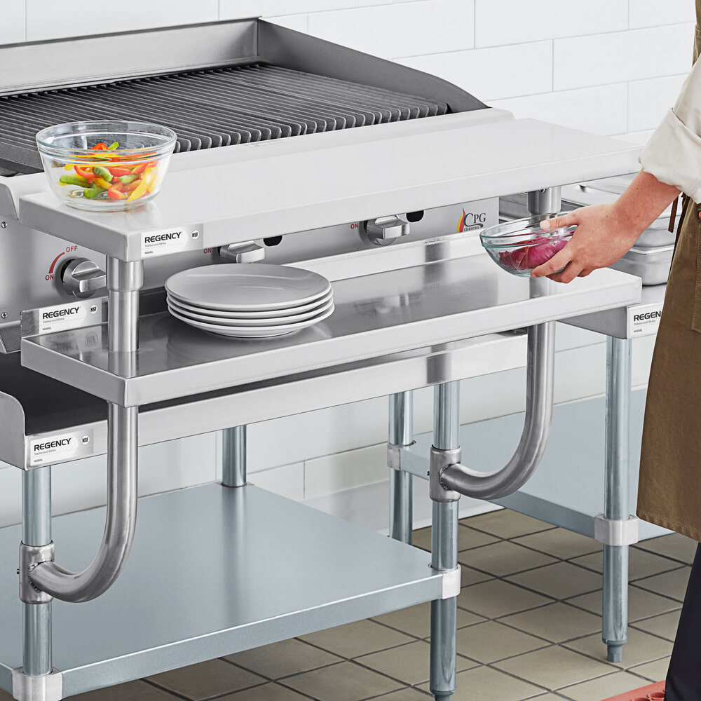 A stainless steel plate shelf mounted above a commercial kitchen equipment stand, holding a stack of plates and a glass bowl with vegetables.