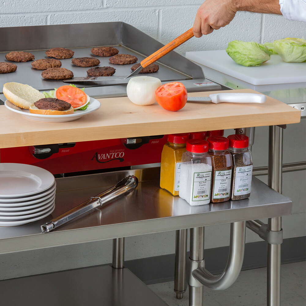 A kitchen scene showing a stainless steel equipment stand with a wooden shelf, various spices, plates, and a griddle with hamburger patties being cooked.