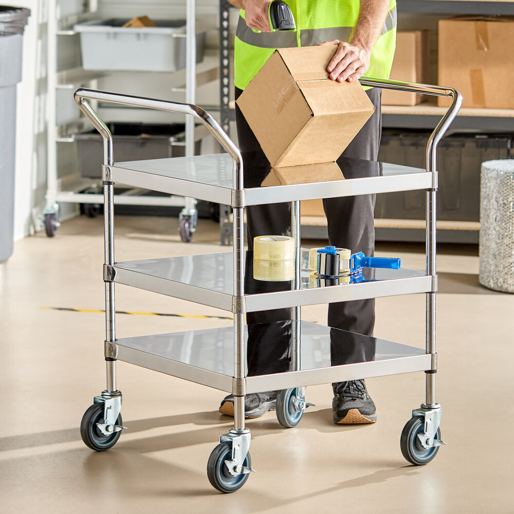 A three-shelf stainless steel utility cart with large swivel casters, shown in a warehouse setting with packing supplies and a person handling a cardboard box on the top shelf.